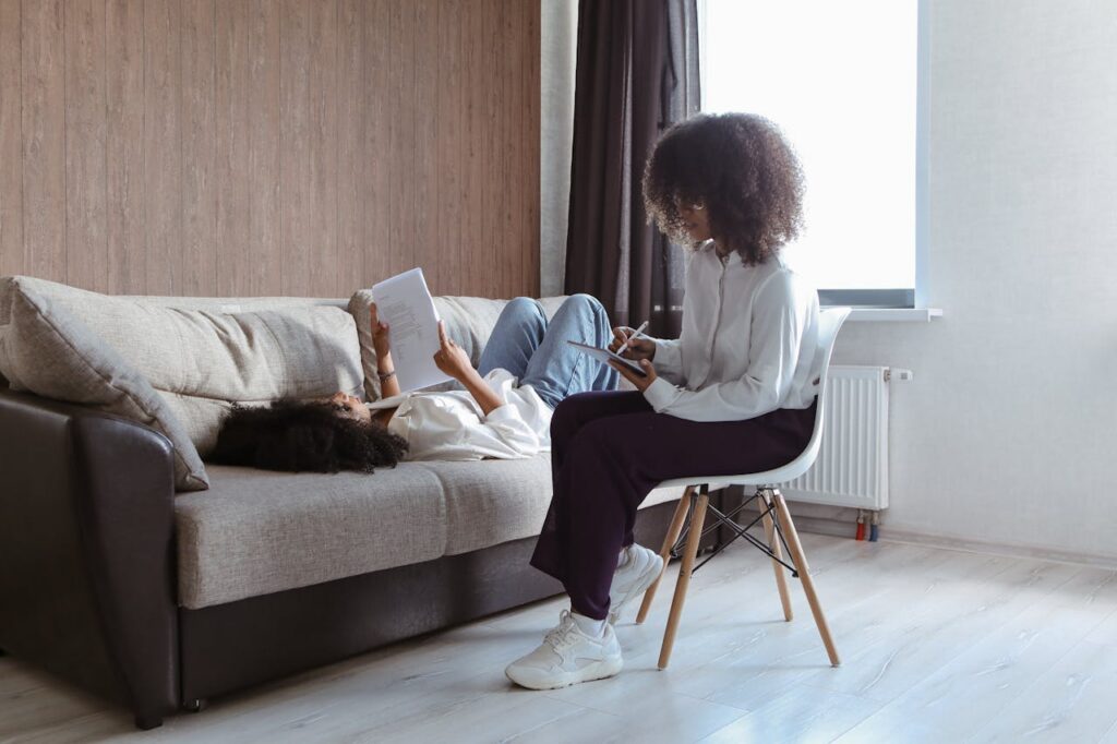 Two Afro-haired women engage in a therapy session seated indoors, fostering communication and wellbeing.
